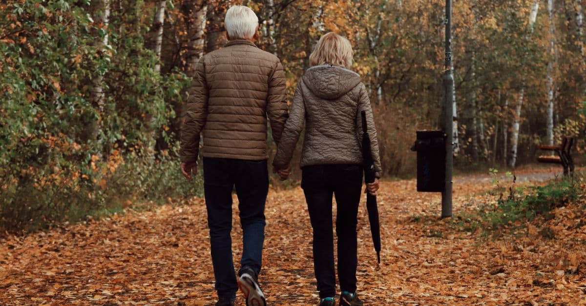 Older couple holding hands while walking a leaf-covered park path in autumn