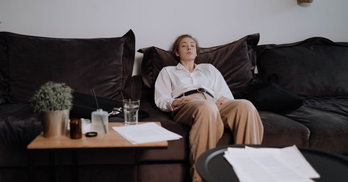 A woman slumped back on a dark sofa with her eyes closed, work papers and a glass of water on the table beside her