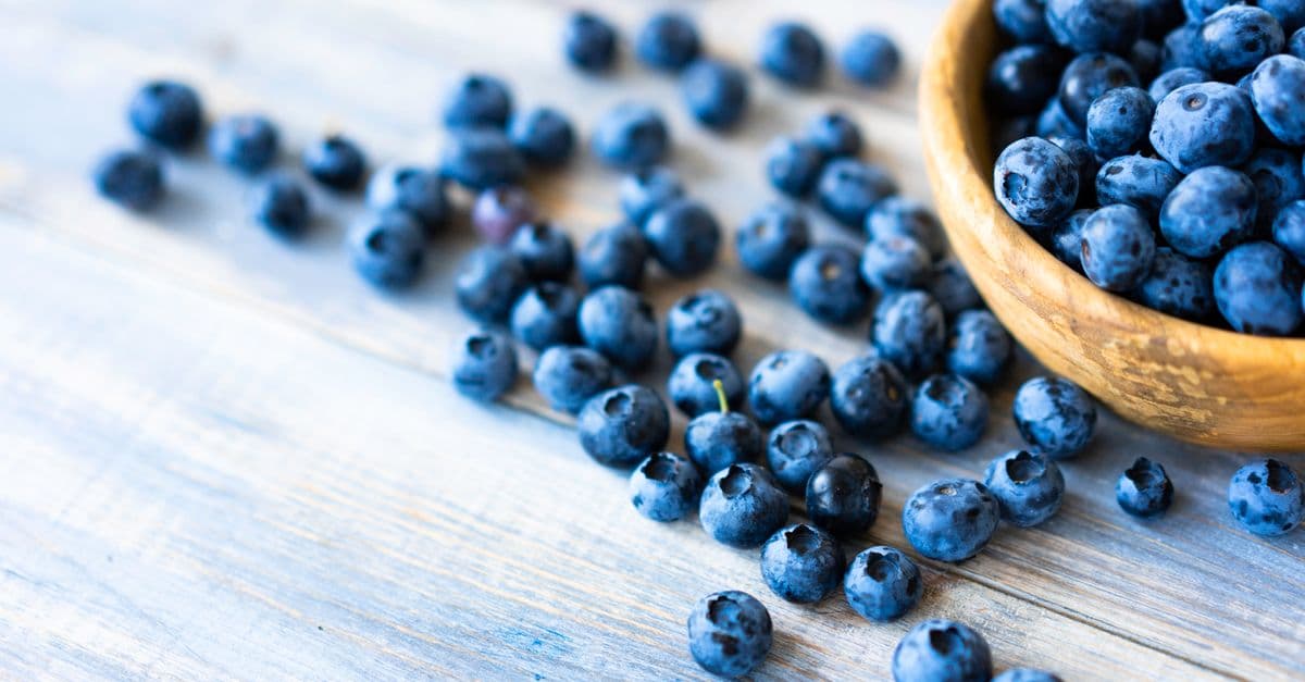 Fresh blueberries spilling from a wooden bowl onto a pale weathered wood table
