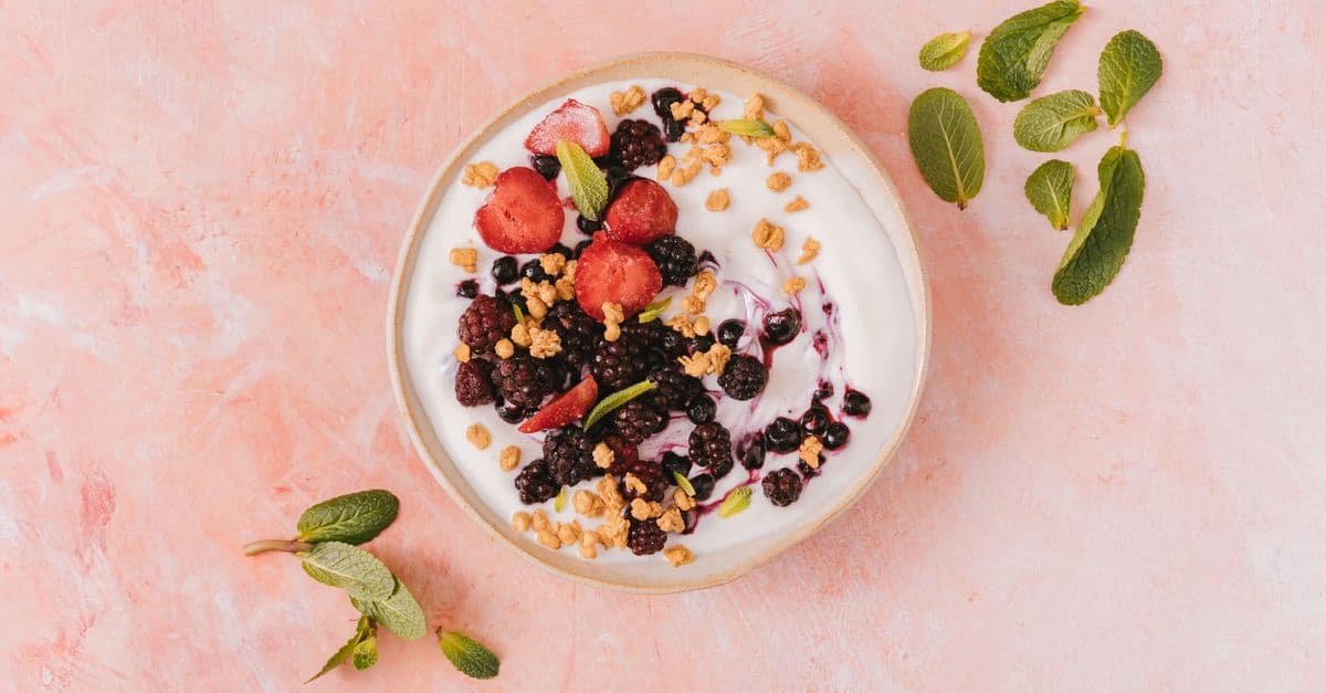 Overhead view of a bowl of plain yogurt topped with granola, blackberries, blueberries and sliced strawberries beside sprigs of mint
