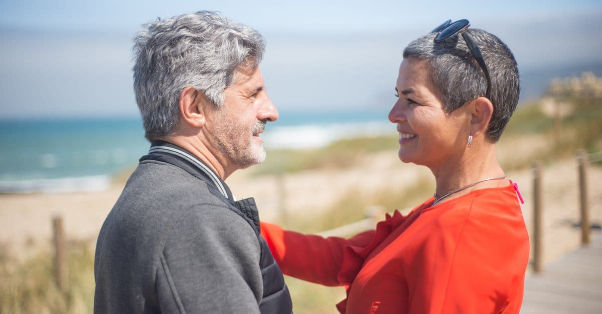 Mature couple standing close together on a sunny beach boardwalk, smiling at one another