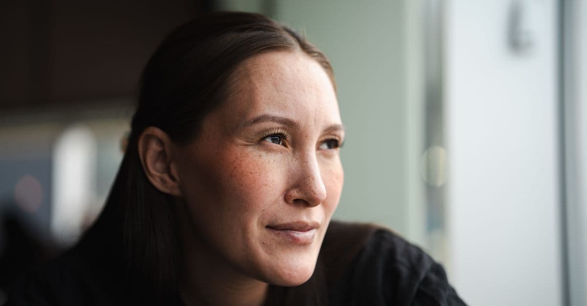A woman with dark hair sits in soft window light, looking calmly out of frame with a faint smile