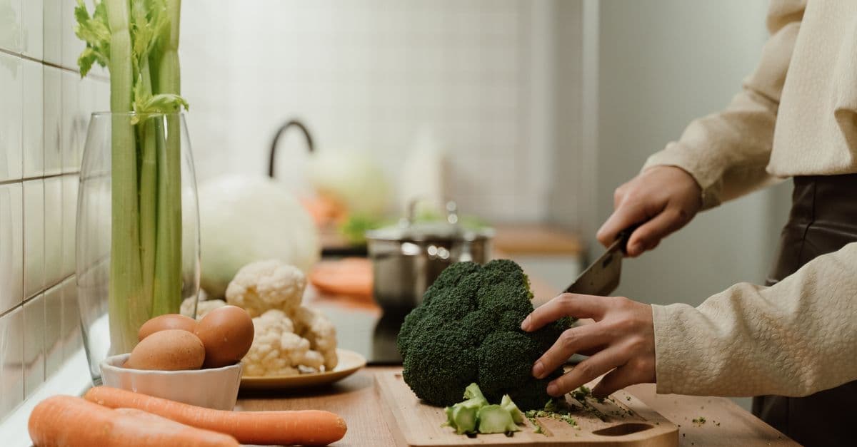 Hands chopping a head of broccoli on a wooden board in a home kitchen, with carrots, eggs, cauliflower and celery nearby