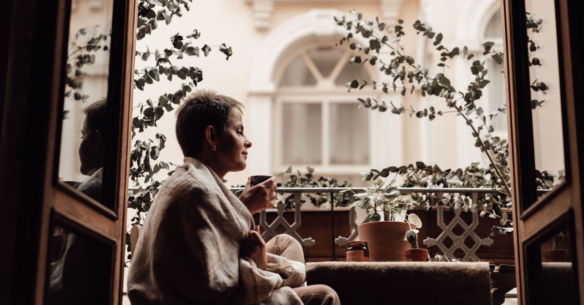 Woman wrapped in a soft blanket sitting on a plant-filled balcony holding a warm cup of tea