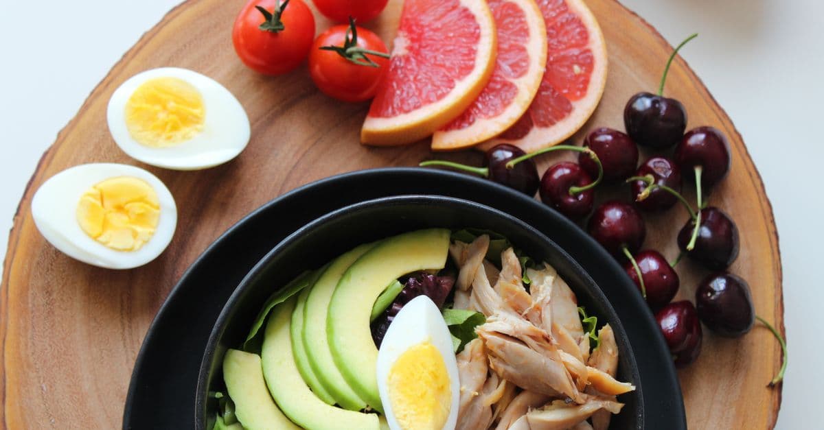 Overhead view of a wooden board with a bowl of salad, sliced avocado and chicken, hard-boiled eggs, cherry tomatoes, grapefruit slices and cherries