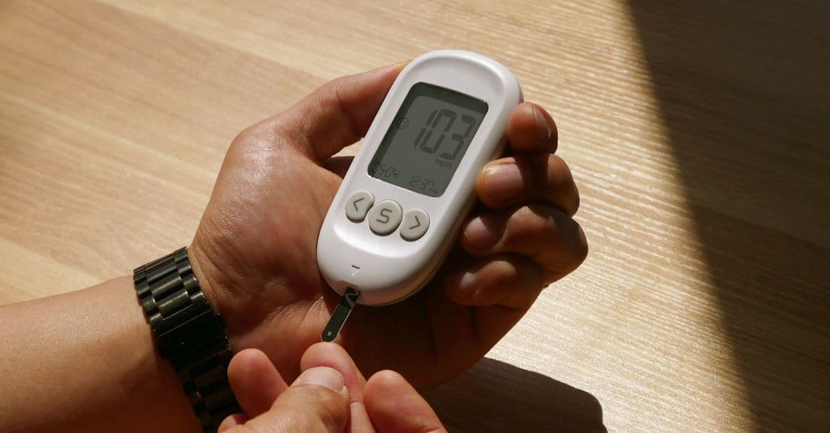 Close-up of hands holding a white blood glucose meter with a test strip inserted, resting over a wooden table