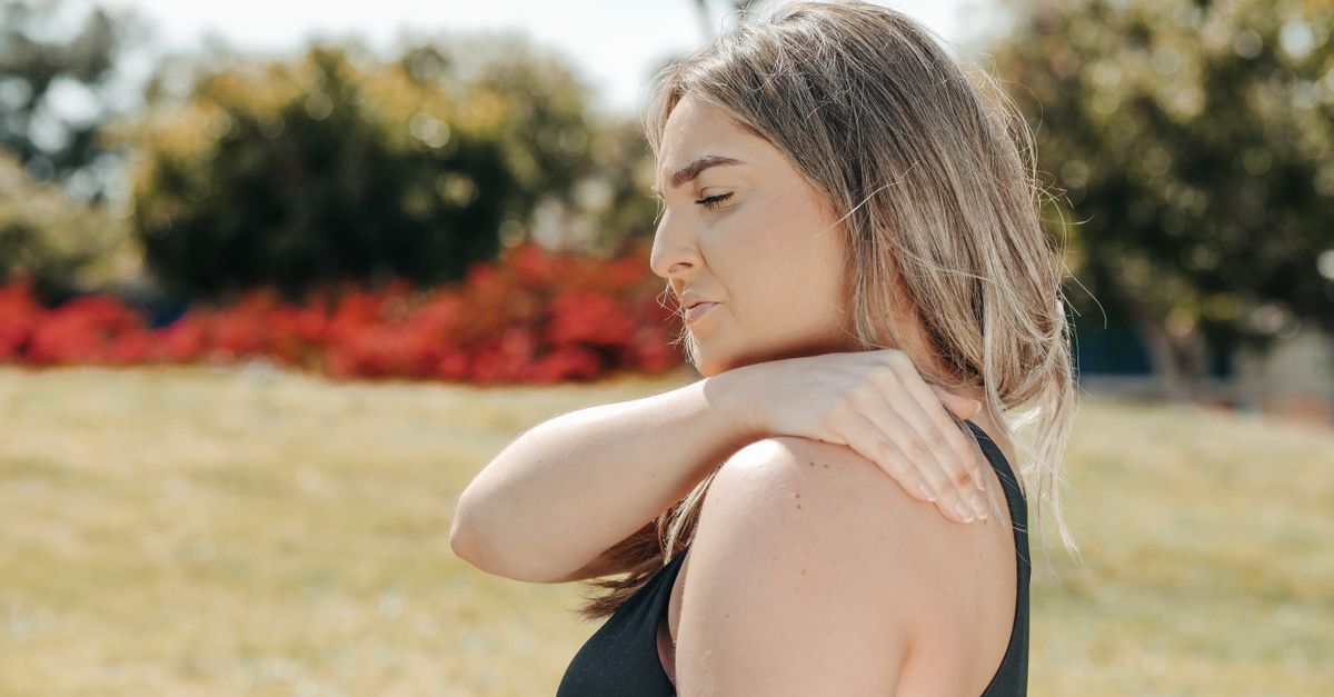 Woman outdoors in a park reaching across to hold her shoulder, eyes closed in discomfort