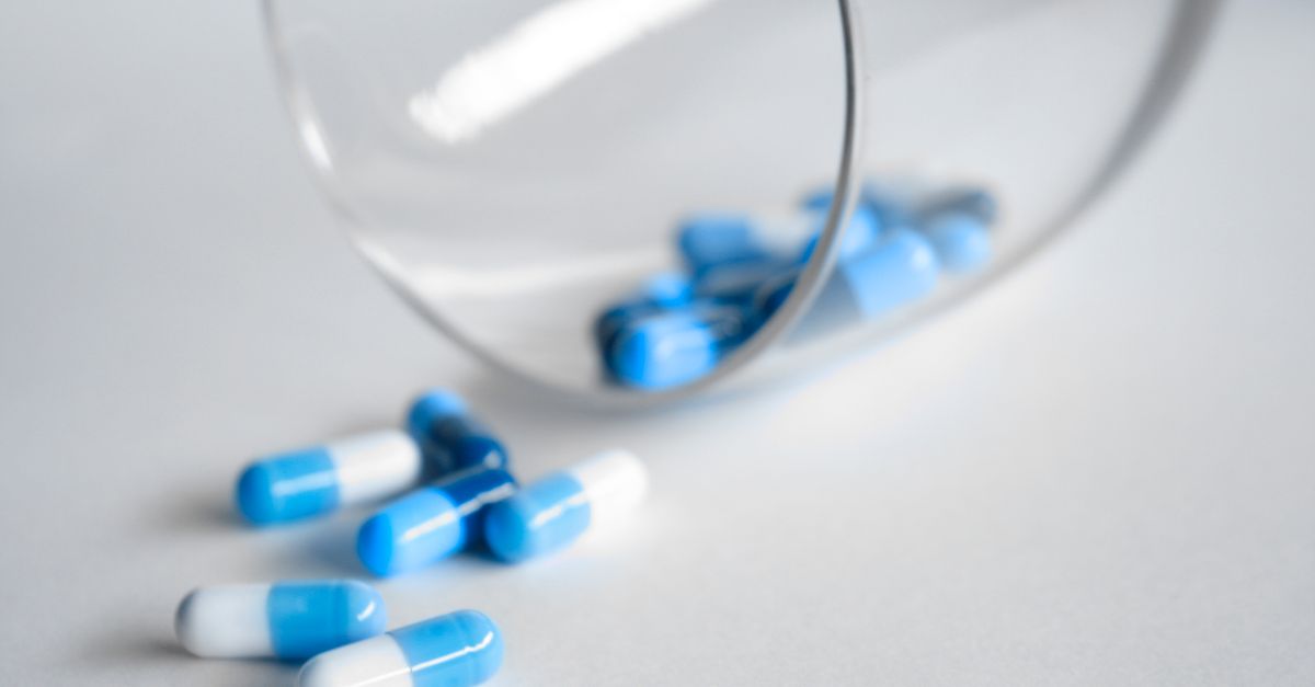 Blue and white two-tone medication capsules spilling from a tipped clear glass onto a clean white surface