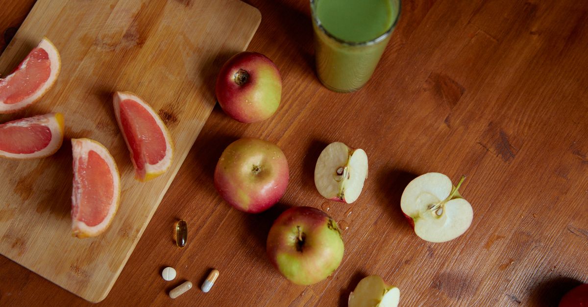 Overhead view of a warm wooden table with sliced pink grapefruit on a cutting board, whole and halved apples, a green vegetable juice, and a few supplement capsules and tablets