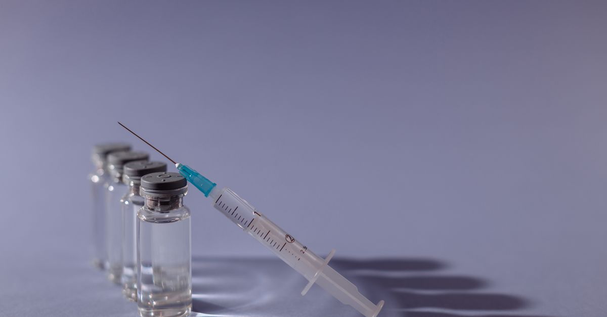 A row of small clear glass medication vials with gray rubber-topped caps beside a fine-gauge syringe resting against them on a smooth pale surface