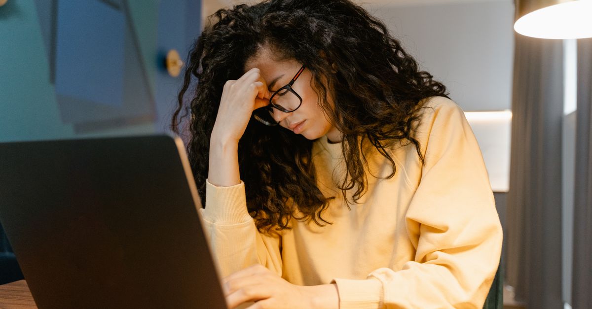 A woman in a yellow sweatshirt and glasses sits at a laptop in the evening, pressing a hand to her forehead with her eyes closed