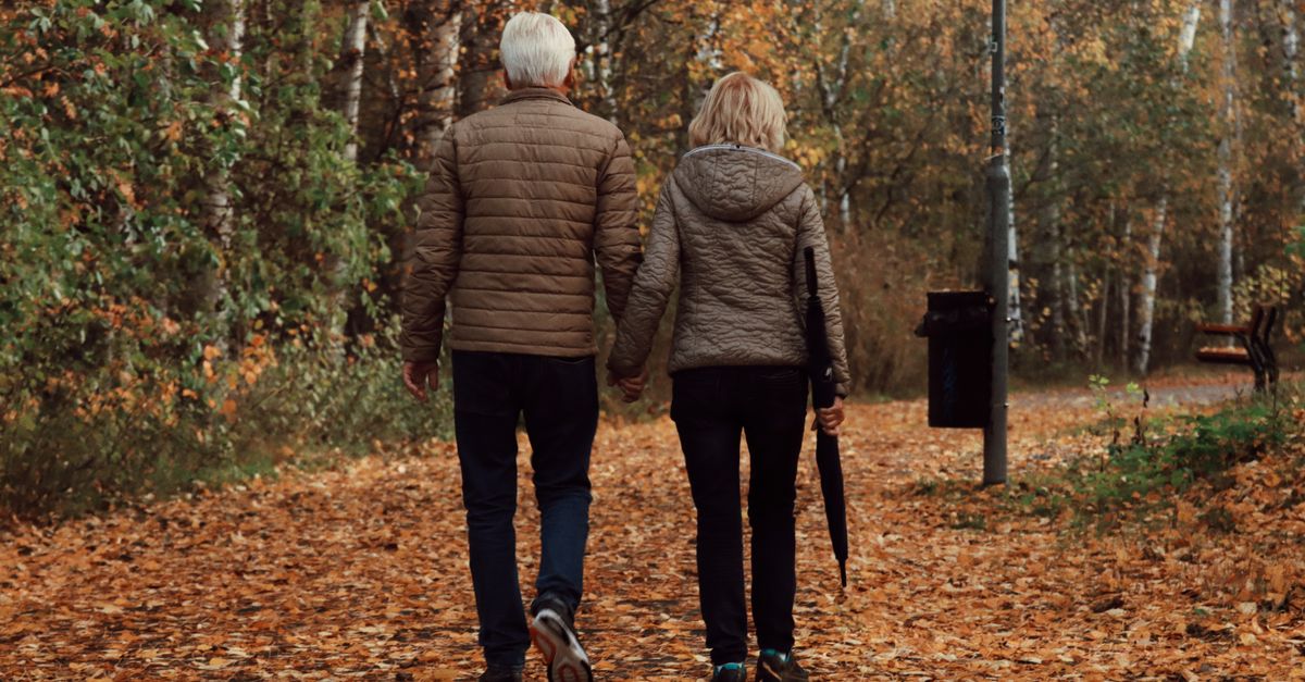 Older couple holding hands while walking a leaf-covered park path in autumn