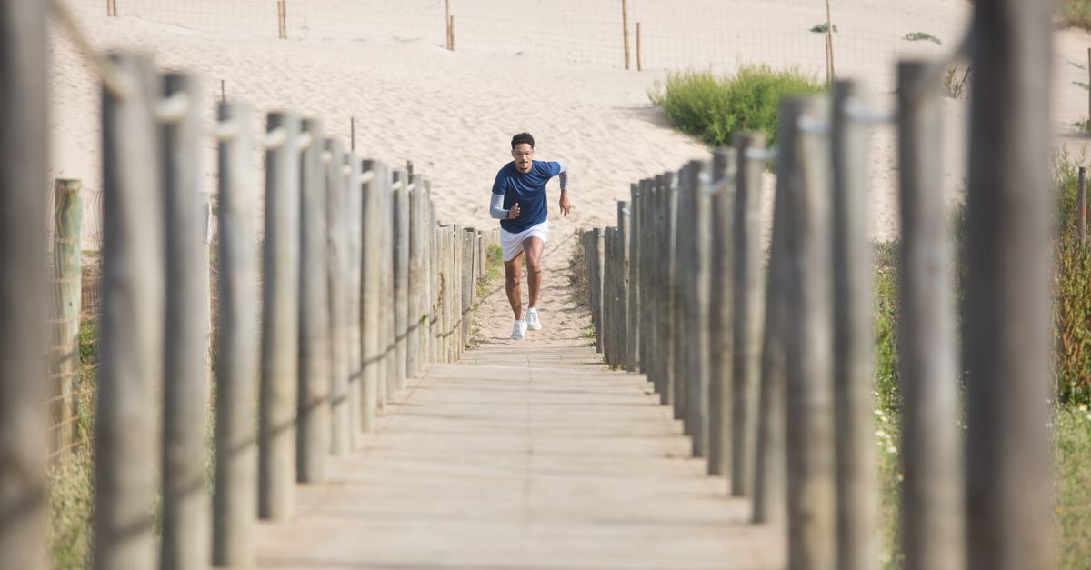 A man in athletic wear runs up a wooden beach boardwalk between weathered dune fence posts on a bright day