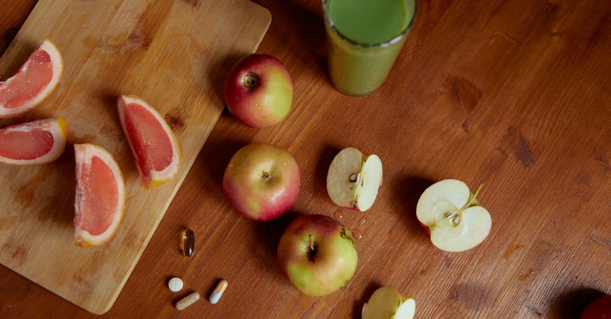 Overhead view of a warm wooden table with sliced pink grapefruit on a cutting board, whole and halved apples, a green vegetable juice, and a few supplement capsules and tablets