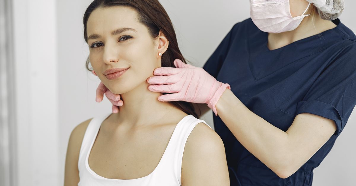 A masked clinician in navy scrubs and gloves palpates the front of a seated woman's neck during a thyroid exam